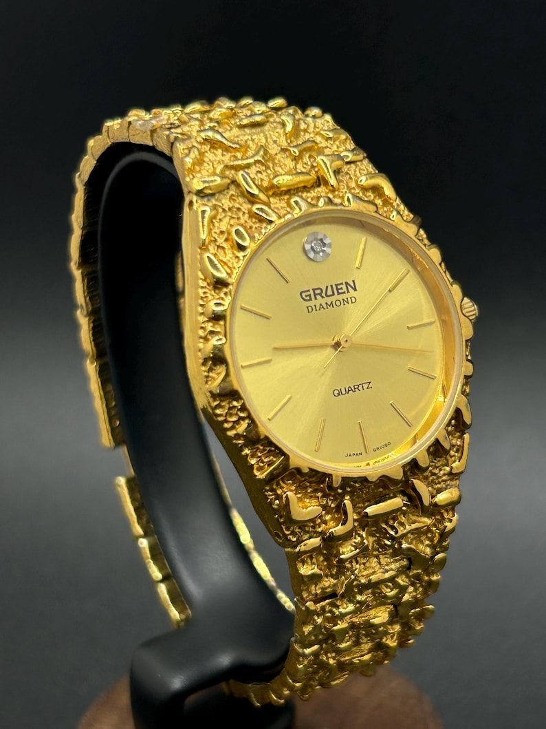 May include: A gold-tone Gruen Diamond quartz watch featuring a textured, nugget-style link bracelet and a round gold dial. The watch face displays the text Gruen Diamond and Quartz, with a single diamond marker at the twelve o'clock position.