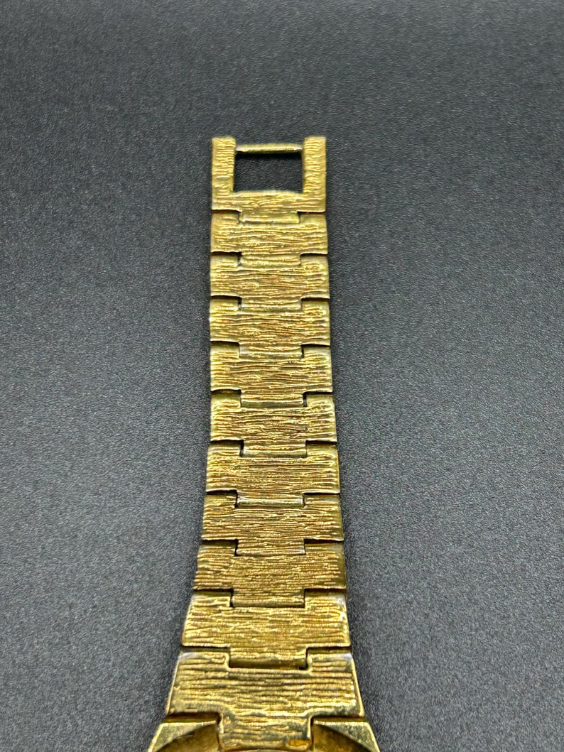 May include: A gold-toned metal watch band featuring a textured, bark-like finish on each rectangular link. The band is composed of multiple interlocking segments that taper slightly toward the top, ending in a simple rectangular clasp connector.
