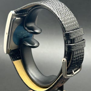K&ouml;nnte beinhalten: Eine rechteckige silberne Armbanduhr mit einem schwarzen, strukturierten Lederarmband. Die Uhr wird auf einem schwarzen St&auml;nder mit Holzsockel pr&auml;sentiert. Das Uhrenarmband hat eine silberne Schnalle.