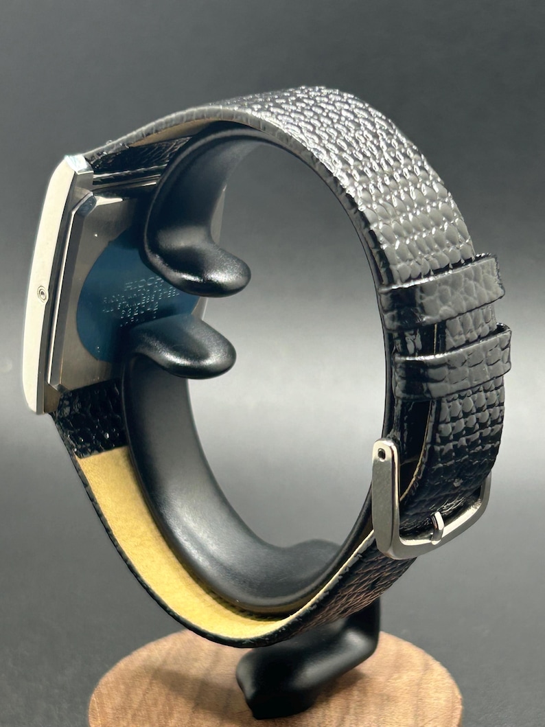 K&ouml;nnte beinhalten: Eine rechteckige silberne Armbanduhr mit einem schwarzen, strukturierten Lederarmband. Die Uhr wird auf einem schwarzen St&auml;nder mit Holzsockel pr&auml;sentiert. Das Uhrenarmband hat eine silberne Schnalle.