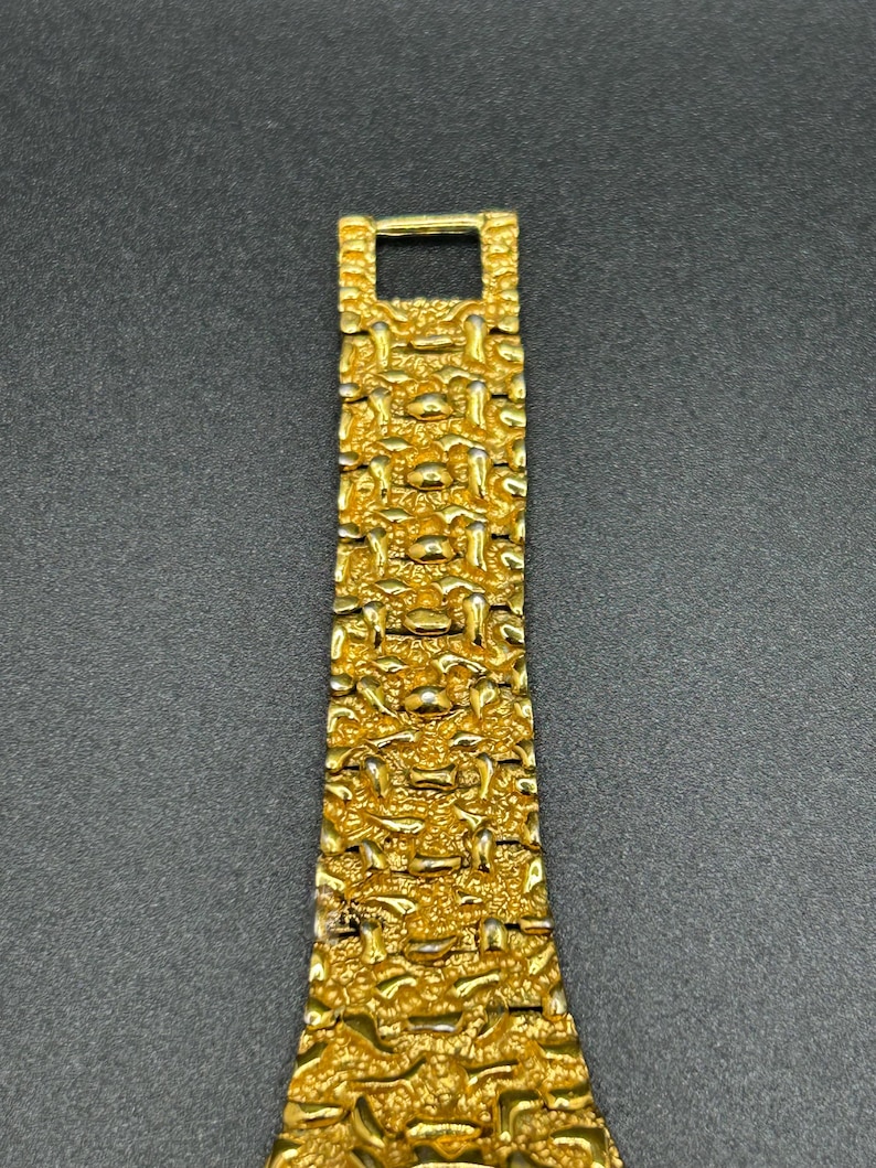 May include: A gold-toned watch band featuring a textured, nugget-style link design. The metal strap consists of multiple interconnected segments with a rough, uneven surface finish, tapering slightly toward the top where a rectangular clasp is visible.