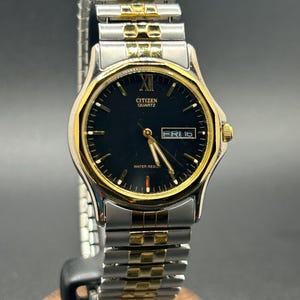 May include: A Citizen Quartz watch with a black dial, gold-tone bezel, and silver and gold-tone band. The watch displays the day and date. The watch is water resistant.