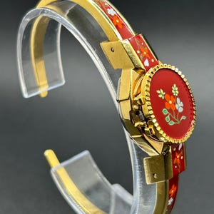 Puede incluir: Una pulsera de brazalete dorada con un dise&ntilde;o floral de esmalte rojo. La pulsera presenta una pieza central redonda y roja con un motivo floral y se exhibe sobre un soporte transparente. El dise&ntilde;o de la pulsera incluye flores naranjas y blancas.