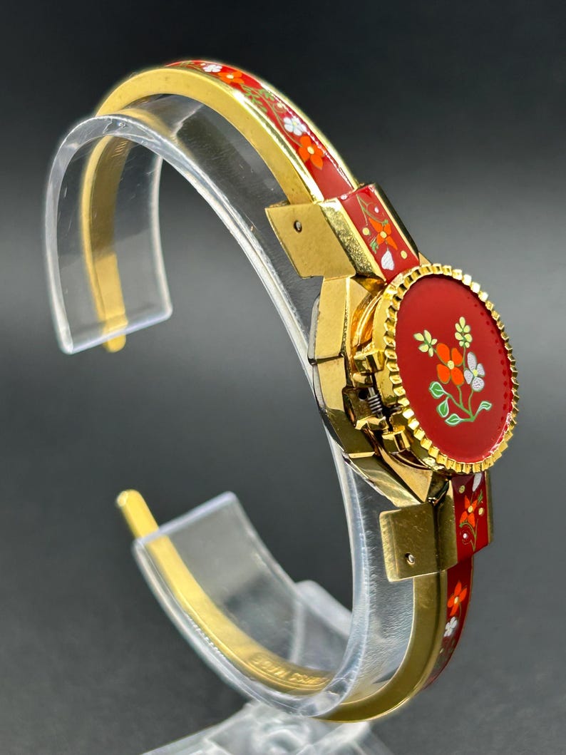 Puede incluir: Una pulsera de brazalete dorada con un dise&ntilde;o floral de esmalte rojo. La pulsera presenta una pieza central redonda y roja con un motivo floral y se exhibe sobre un soporte transparente. El dise&ntilde;o de la pulsera incluye flores naranjas y blancas.