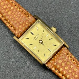 May include: A gold-toned Caravelle Quartz watch with a rectangular face and a brown leather band. The watch face has black markers and hands. The band is a textured, light brown color.