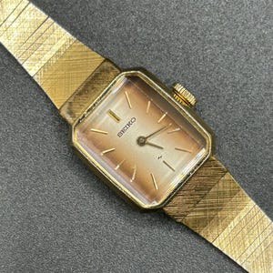 May include: A vintage Seiko watch with a gold-toned rectangular face and matching strap. The watch face has gold markers and hands, with the Seiko logo. The strap is a textured, flexible design. A classic timepiece.