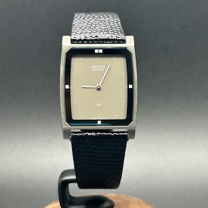 K&ouml;nnte beinhalten: Eine Vintage Ricoh Quartz-Uhr mit einem rechteckigen, silberfarbenen Geh&auml;use und einem hellbeigen Zifferblatt. Die Uhr hat ein schwarzes Lederarmband und wird auf einem Holzst&auml;nder pr&auml;sentiert. Das Zifferblatt zeigt den Markennamen.