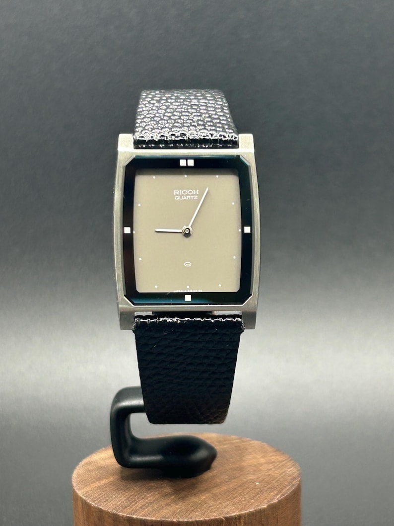 K&ouml;nnte beinhalten: Eine Vintage Ricoh Quartz-Uhr mit einem rechteckigen, silberfarbenen Geh&auml;use und einem hellbeigen Zifferblatt. Die Uhr hat ein schwarzes Lederarmband und wird auf einem Holzst&auml;nder pr&auml;sentiert. Das Zifferblatt zeigt den Markennamen.