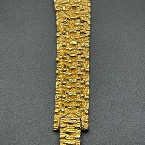 May include: A gold-toned watch bracelet featuring a highly textured, nugget-like surface pattern. The metal links are interconnected in a repeating, irregular design that creates a rugged, three-dimensional appearance across the entire length.