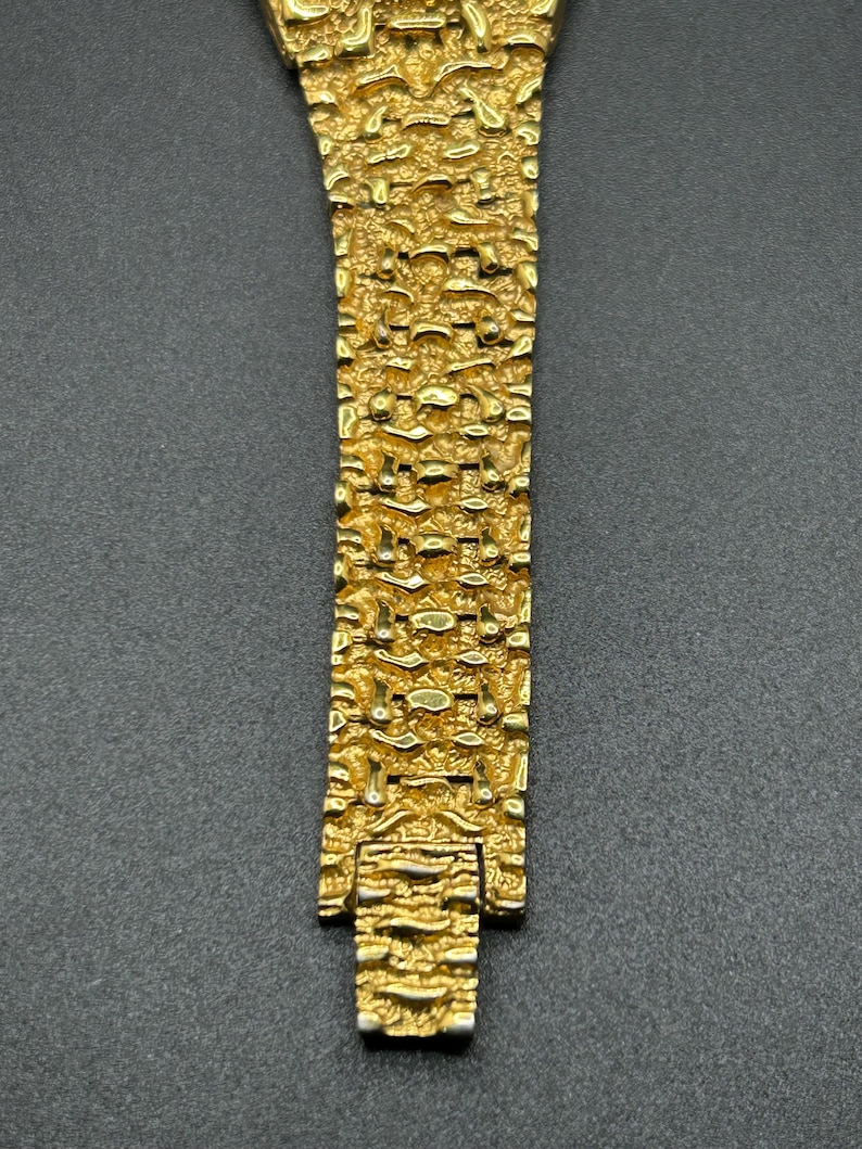 May include: A gold-toned watch bracelet featuring a highly textured, nugget-like surface pattern. The metal links are interconnected in a repeating, irregular design that creates a rugged, three-dimensional appearance across the entire length.