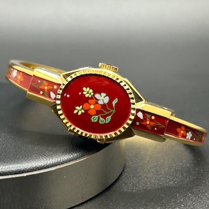 Puede incluir: Una pulsera dorada con una esfera y una banda de esmalte rojo. La esfera presenta un dise&ntilde;o floral con detalles en blanco, naranja y verde. La banda es roja con motivos florales. La pulsera es de estilo articulado.