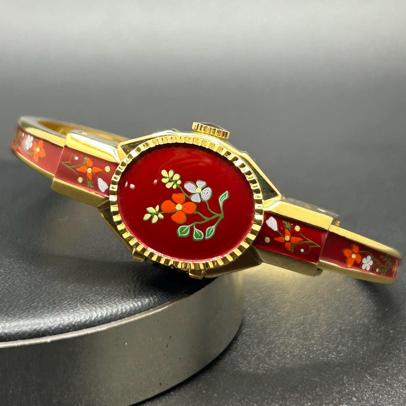 Puede incluir: Una pulsera dorada con una esfera y una banda de esmalte rojo. La esfera presenta un dise&ntilde;o floral con detalles en blanco, naranja y verde. La banda es roja con motivos florales. La pulsera es de estilo articulado.