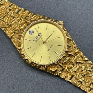 May include: A gold-tone quartz wristwatch featuring a textured nugget-style link band and a round gold dial. The dial displays the text GRUEN DIAMOND and QUARTZ, with a single diamond marker at the twelve o'clock position and thin gold hands.
