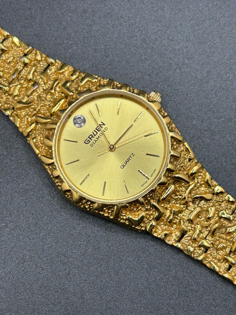 May include: A gold-tone quartz wristwatch featuring a textured nugget-style link band and a round gold dial. The dial displays the text GRUEN DIAMOND and QUARTZ, with a single diamond marker at the twelve o'clock position and thin gold hands.