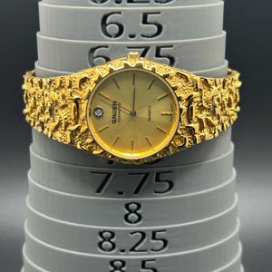 May include: A gold-tone Gruen Diamond quartz watch with a textured nugget-style band. The watch face features a single diamond marker at the 12 o'clock position. It is displayed on a gray sizing mandrel showing a circumference of 7.5 inches.