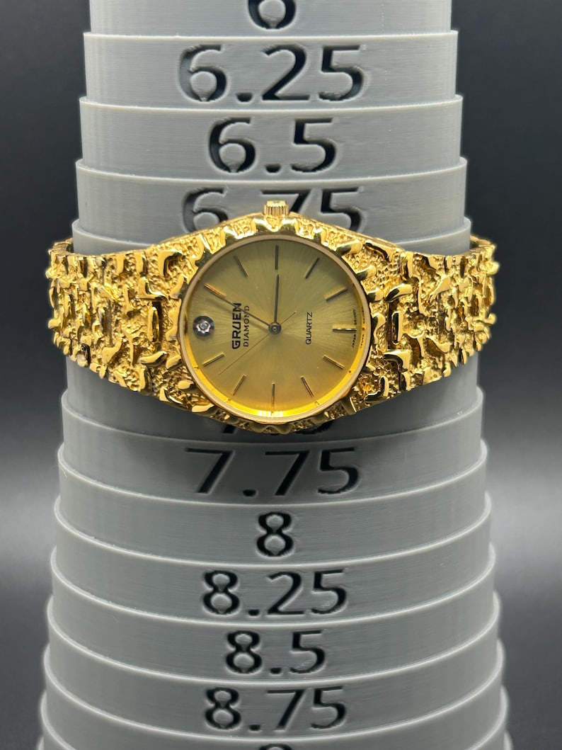 May include: A gold-tone Gruen Diamond quartz watch with a textured nugget-style band. The watch face features a single diamond marker at the 12 o'clock position. It is displayed on a gray sizing mandrel showing a circumference of 7.5 inches.