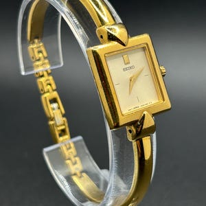 May include: A gold-toned Seiko wristwatch with a rectangular face and a cream-colored dial. The watch features a gold band and a minimalist design. The watch is displayed on a clear stand.