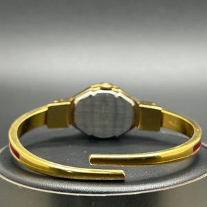 Puede incluir: Reloj de pulsera dorado con un detalle de raya roja. El reloj presenta una correa dorada y una esfera plateada. El reloj se muestra sobre una superficie negra. El reloj es un accesorio elegante.