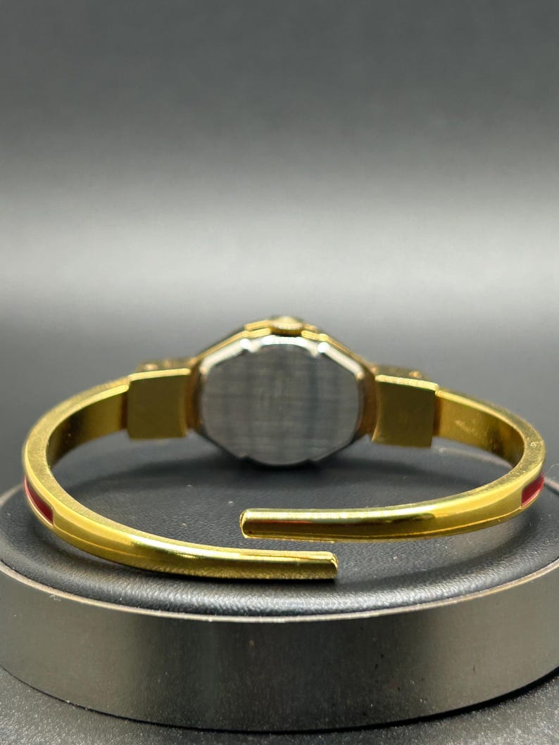 Puede incluir: Reloj de pulsera dorado con un detalle de raya roja. El reloj presenta una correa dorada y una esfera plateada. El reloj se muestra sobre una superficie negra. El reloj es un accesorio elegante.