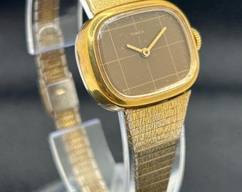Reloj Timex vintage para mujer, de cuerda manual, con esfera cuadrada dorada y correa texturizada.