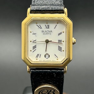 May include: A gold-toned Bulova Quartz watch with a rectangular face and black leather band. The watch face is white with black Roman numerals and gold hands. The date is displayed at the 3 o'clock position. The band has a gold emblem with the letters "CB".