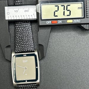 K&ouml;nnte beinhalten: Eine silberfarbene Ricoh Quartz-Uhr mit rechteckigem Zifferblatt und schwarzem, strukturiertem Armband. Das Zifferblatt ist hellbraun mit silbernen Zeigern und dem Ricoh Quartz-Logo. Die Uhr misst 2,75 cm.
