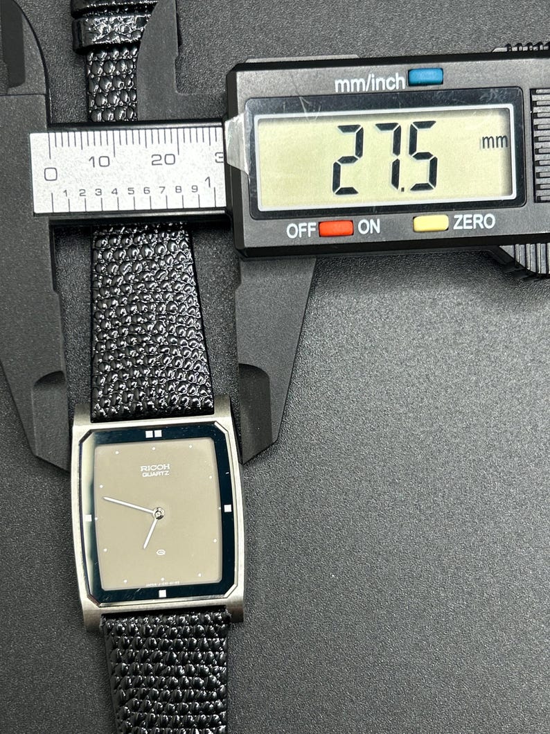 K&ouml;nnte beinhalten: Eine silberfarbene Ricoh Quartz-Uhr mit rechteckigem Zifferblatt und schwarzem, strukturiertem Armband. Das Zifferblatt ist hellbraun mit silbernen Zeigern und dem Ricoh Quartz-Logo. Die Uhr misst 2,75 cm.