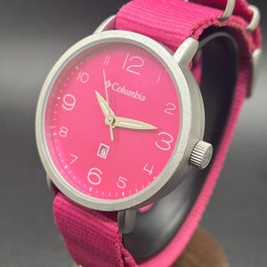 May include: A Columbia watch with a bright pink face and matching pink fabric band. The watch has a silver-colored bezel and hands, with the date displayed at the 6 o'clock position. The watch is displayed on a wooden stand.