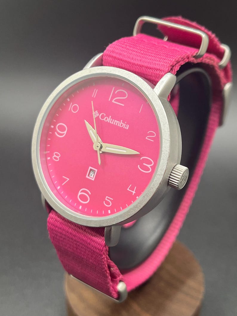May include: A Columbia watch with a bright pink face and matching pink fabric band. The watch has a silver-colored bezel and hands, with the date displayed at the 6 o'clock position. The watch is displayed on a wooden stand.