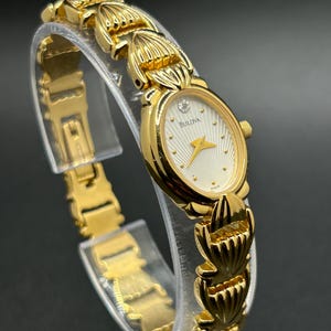 Peut inclure: Une montre-bracelet Bulova dorée dotée d'un boîtier ovale avec un cadran blanc texturé, des aiguilles dorées et un diamant unique à la position douze heures. La montre possède un bracelet à maillons décoratifs au motif en forme de coquille.
