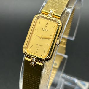 Peut inclure: Une montre à quartz Lassale Seiko dorée avec un cadran rectangulaire et un bracelet en maille assorti. La montre présente un cadran doré avec des aiguilles et des marqueurs noirs, et de petits accents de diamants. La montre est exposée sur un support transparent.