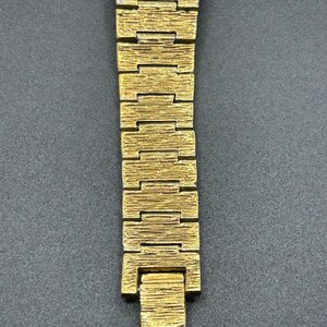 May include: A gold-toned metal watch bracelet featuring a textured, bark-like finish on each rectangular link. The band consists of multiple interlocking segments that taper slightly toward the bottom, ending in a flat, rectangular clasp component.