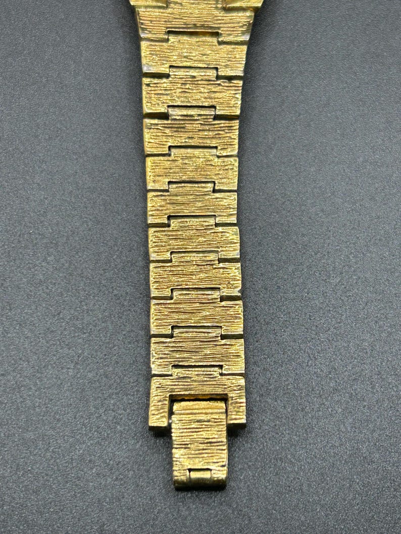 May include: A gold-toned metal watch bracelet featuring a textured, bark-like finish on each rectangular link. The band consists of multiple interlocking segments that taper slightly toward the bottom, ending in a flat, rectangular clasp component.