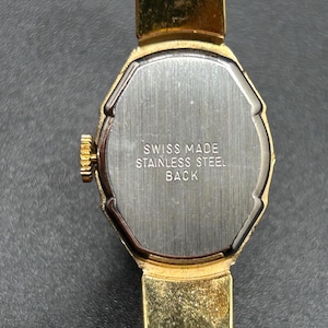 Puede incluir: Primer plano de un reloj de pulsera vintage dorado. El reloj tiene una esfera rectangular con una parte trasera plateada. El texto "SWISS MADE STAINLESS STEEL BACK" es visible. La correa del reloj tambi&eacute;n es dorada.