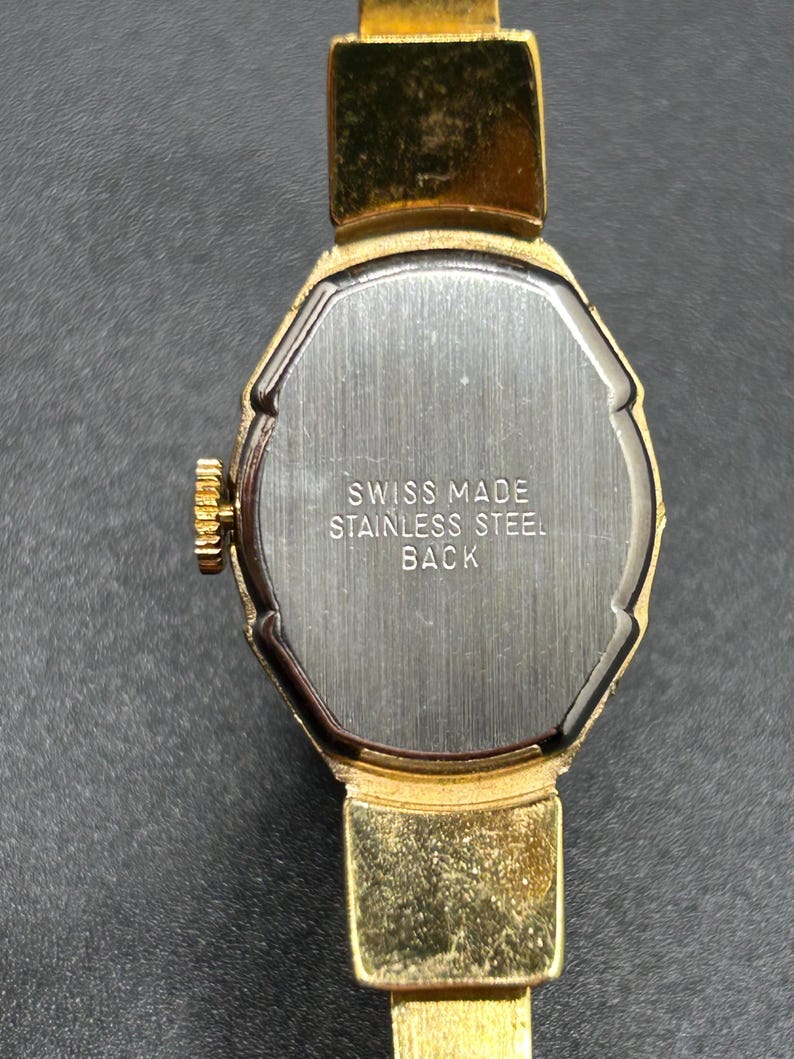 Puede incluir: Primer plano de un reloj de pulsera vintage dorado. El reloj tiene una esfera rectangular con una parte trasera plateada. El texto "SWISS MADE STAINLESS STEEL BACK" es visible. La correa del reloj tambi&eacute;n es dorada.