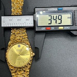May include: A gold-tone Gruen Diamond quartz watch with a textured nugget-style link band. The watch face features a single diamond marker at the twelve o'clock position. A digital caliper displays a measurement of 34.9 mm across the watch case.