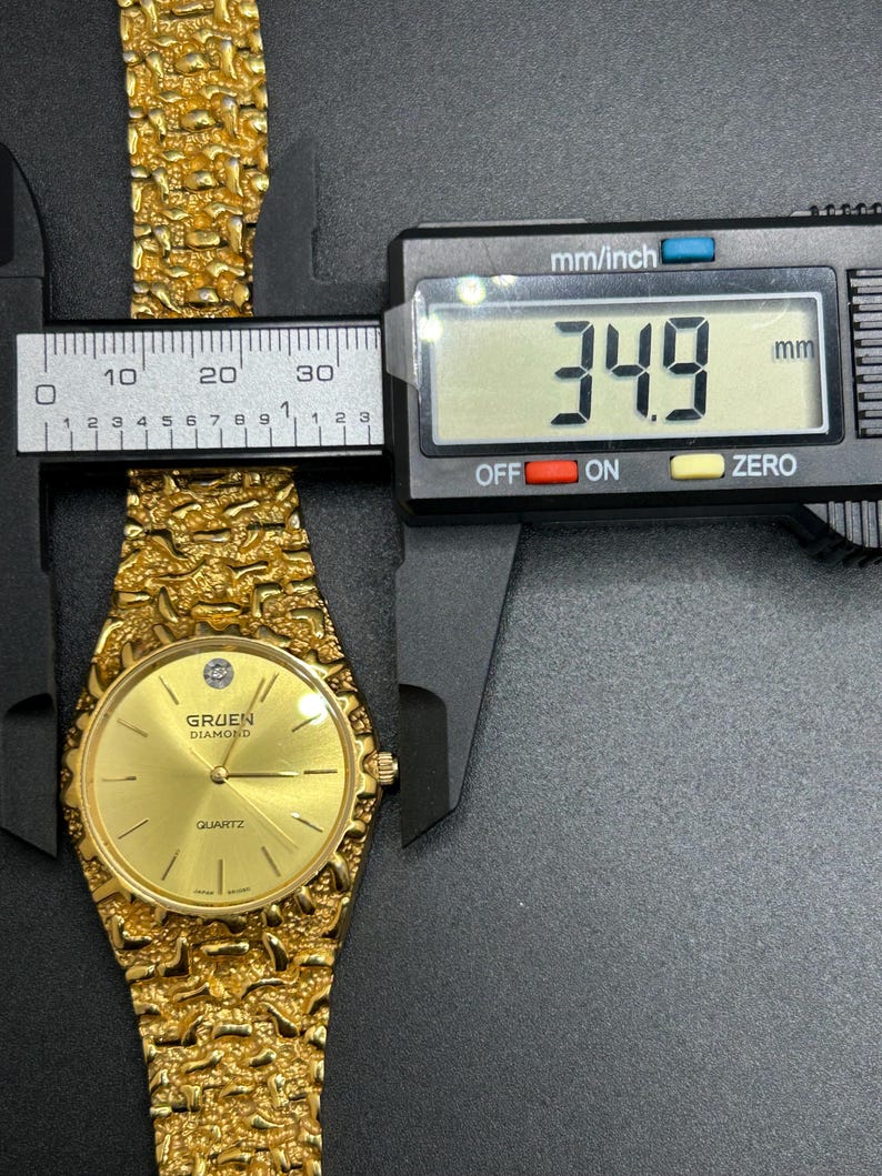 May include: A gold-tone Gruen Diamond quartz watch with a textured nugget-style link band. The watch face features a single diamond marker at the twelve o'clock position. A digital caliper displays a measurement of 34.9 mm across the watch case.