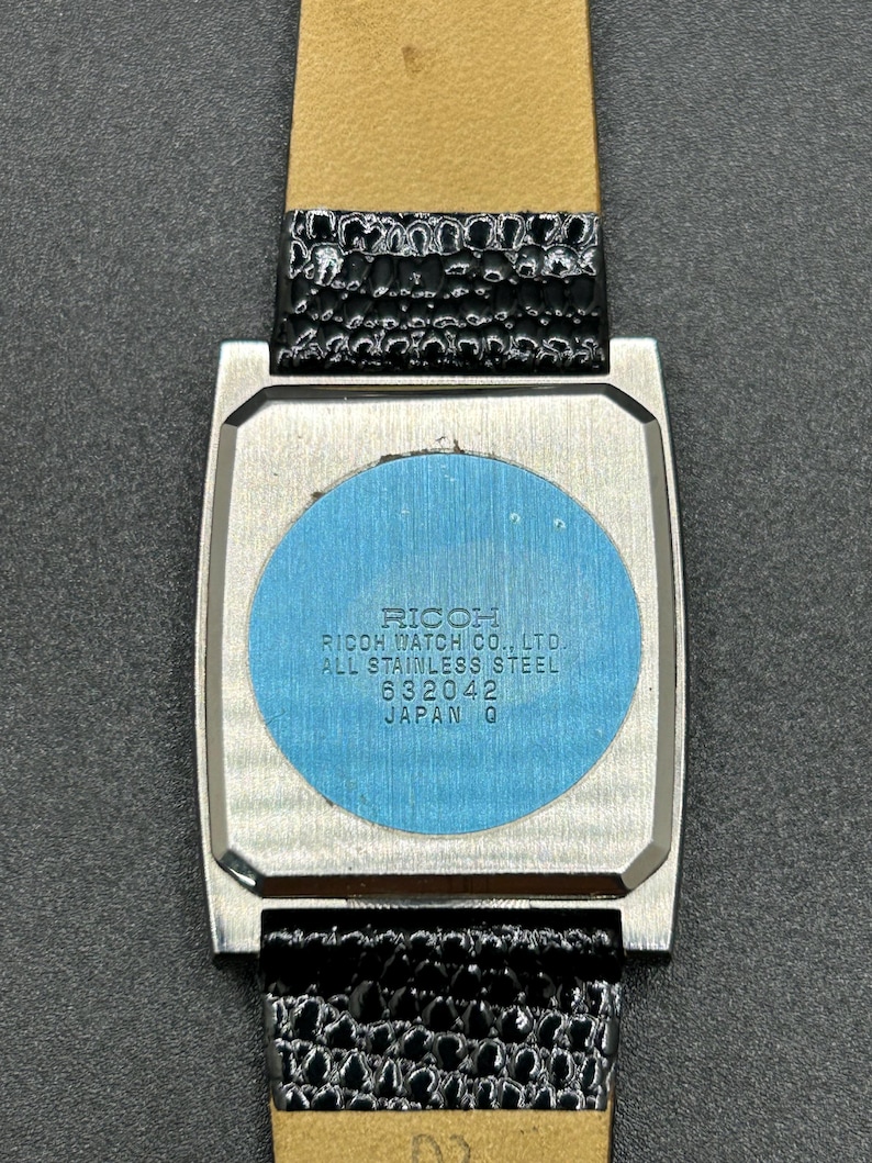K&ouml;nnte beinhalten: Eine Vintage-Ricoh-Uhr mit einem rechteckigen Edelstahlgeh&auml;use und einem schwarzen Lederarmband. Auf der R&uuml;ckseite der Uhr steht unter anderem "Ricoh Watch Co., Ltd., All Stainless Steel, 632042, Japan Q."