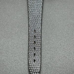 K&ouml;nnte beinhalten: Nahaufnahme eines schwarzen Uhrenarmbands mit strukturierter Oberfl&auml;che. Das Armband hat ein Muster aus kleinen, verbundenen Kreisen. Es gibt mehrere L&ouml;cher zur Anpassung. Der Verschluss ist silberfarben. Das Armband ist f&uuml;r eine Armbanduhr konzipiert.