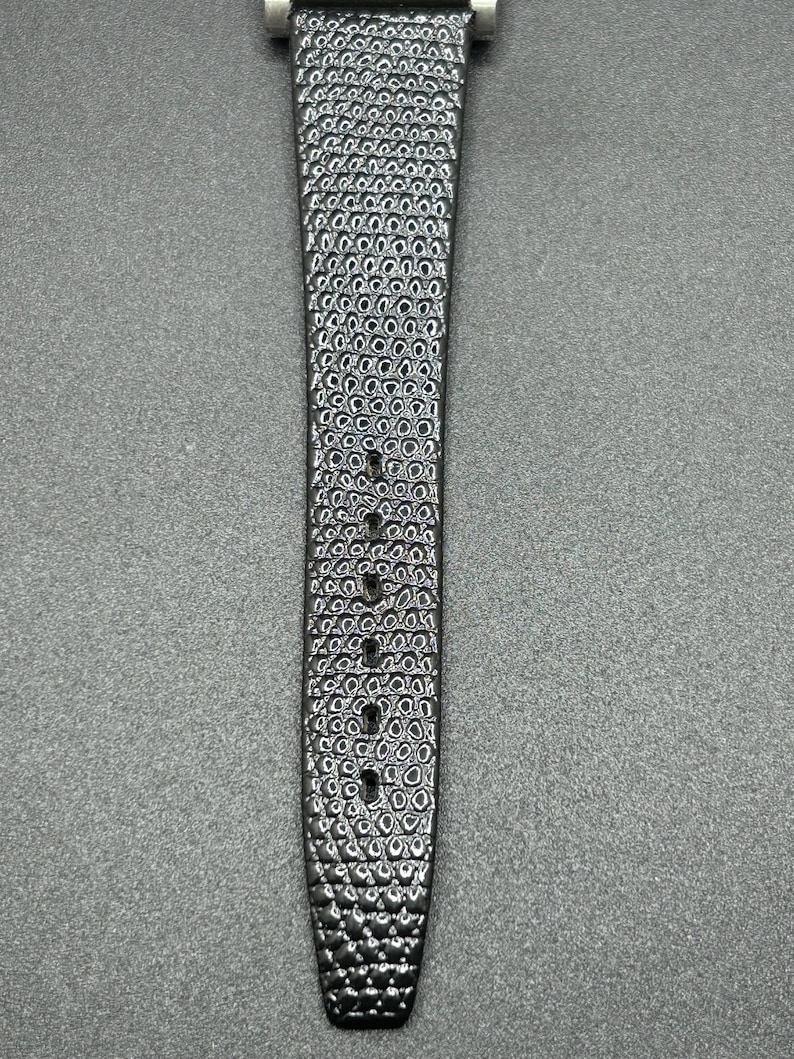 K&ouml;nnte beinhalten: Nahaufnahme eines schwarzen Uhrenarmbands mit strukturierter Oberfl&auml;che. Das Armband hat ein Muster aus kleinen, verbundenen Kreisen. Es gibt mehrere L&ouml;cher zur Anpassung. Der Verschluss ist silberfarben. Das Armband ist f&uuml;r eine Armbanduhr konzipiert.