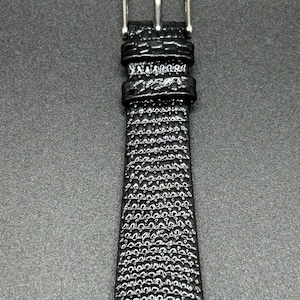 K&ouml;nnte beinhalten: Nahaufnahme eines schwarzen Uhrenarmbands mit einem strukturierten, blasenartigen Muster. Das Armband besteht aus einem gl&auml;nzenden Material und hat eine silberfarbene Schnalle. Das Uhrenarmband ist ein klassisches Design.