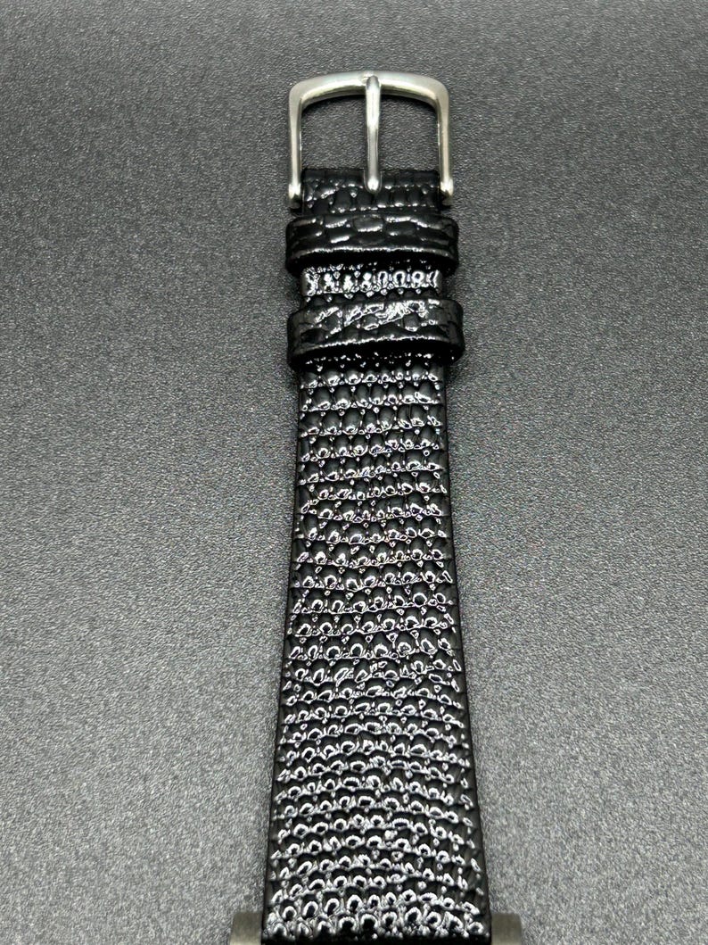 K&ouml;nnte beinhalten: Nahaufnahme eines schwarzen Uhrenarmbands mit einem strukturierten, blasenartigen Muster. Das Armband besteht aus einem gl&auml;nzenden Material und hat eine silberfarbene Schnalle. Das Uhrenarmband ist ein klassisches Design.