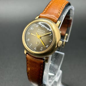 May include: A vintage Bulova Accutron watch with a gold-tone case and a brown leather strap. The watch face is a muted gold color with black numerals and hands. The watch is displayed on a clear stand.