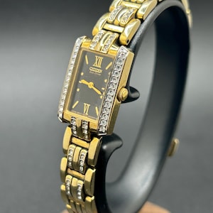 May include: A gold-tone Citizen Eco-Drive rectangular wristwatch with a black dial, gold hands, and Roman numeral markers at twelve and six. The bezel and the links of the metal bracelet are embellished with clear, sparkling crystals.