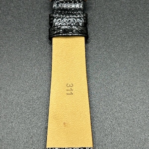K&ouml;nnte beinhalten: Nahaufnahme eines schwarzen Uhrenarmbands mit einer silbernen Schnalle. Das Armband hat eine strukturierte, gl&auml;nzende Oberfl&auml;che. Die Innenseite des Armbands ist beige mit der aufgedruckten Nummer "311". Das Uhrenarmband liegt auf einer dunkelgrauen Oberfl&auml;che.