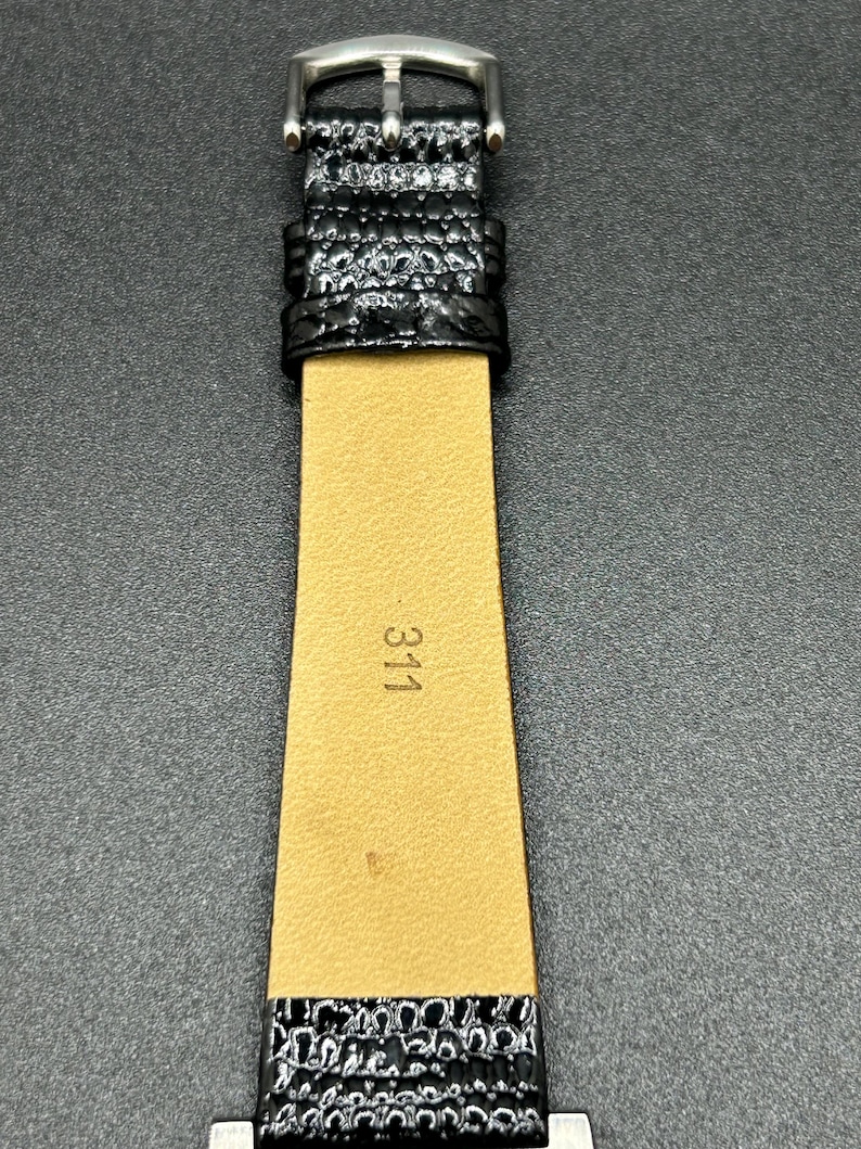 K&ouml;nnte beinhalten: Nahaufnahme eines schwarzen Uhrenarmbands mit einer silbernen Schnalle. Das Armband hat eine strukturierte, gl&auml;nzende Oberfl&auml;che. Die Innenseite des Armbands ist beige mit der aufgedruckten Nummer "311". Das Uhrenarmband liegt auf einer dunkelgrauen Oberfl&auml;che.