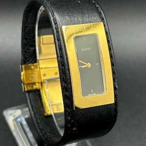 May include: A black leather Gucci watch with a gold rectangular face and a dark gray dial. The watch has a gold clasp and is displayed on a clear stand. The watch face has the word "Gucci" printed on it.
