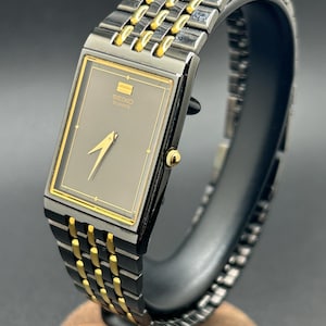 May include: A rectangular Seiko Quartz watch with a gray face and gold hands. The watch has a black and gold band. The watch is displayed on a black stand with a wooden base. The watch face has the Seiko Quartz logo.
