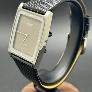 K&ouml;nnte beinhalten: Eine Vintage Ricoh Quartz-Uhr mit rechteckigem Zifferblatt und strukturiertem schwarzem Lederarmband. Das Zifferblatt der Uhr ist hellbraun mit silbernen Zeigern und Markierungen. Die Uhr wird auf einem schwarzen St&auml;nder mit Holzsockel pr&auml;sentiert.