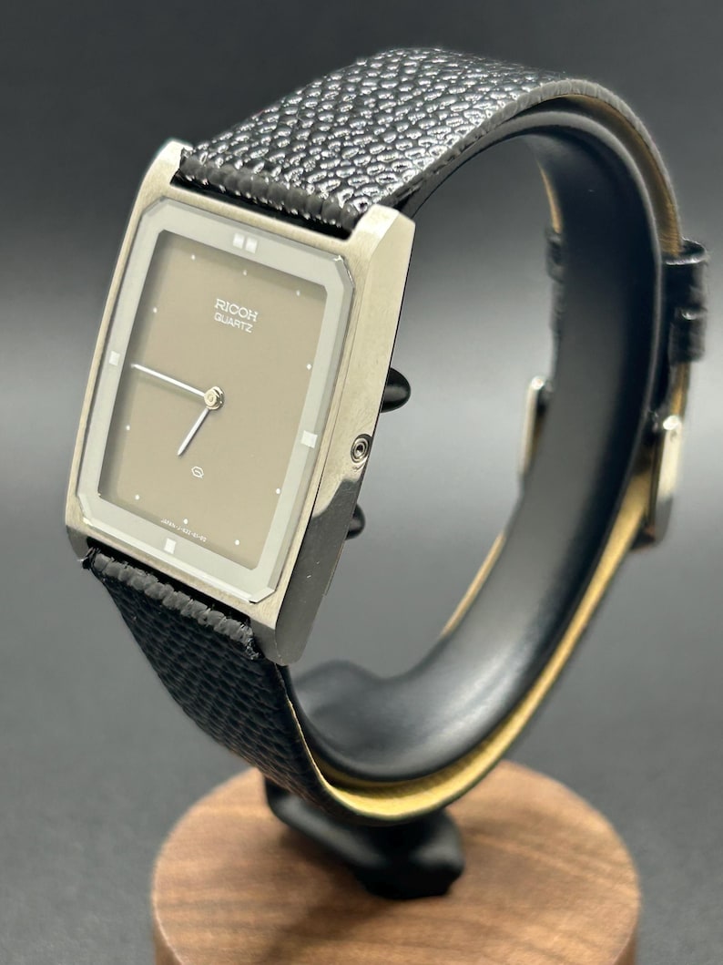 K&ouml;nnte beinhalten: Eine Vintage Ricoh Quartz-Uhr mit rechteckigem Zifferblatt und strukturiertem schwarzem Lederarmband. Das Zifferblatt der Uhr ist hellbraun mit silbernen Zeigern und Markierungen. Die Uhr wird auf einem schwarzen St&auml;nder mit Holzsockel pr&auml;sentiert.